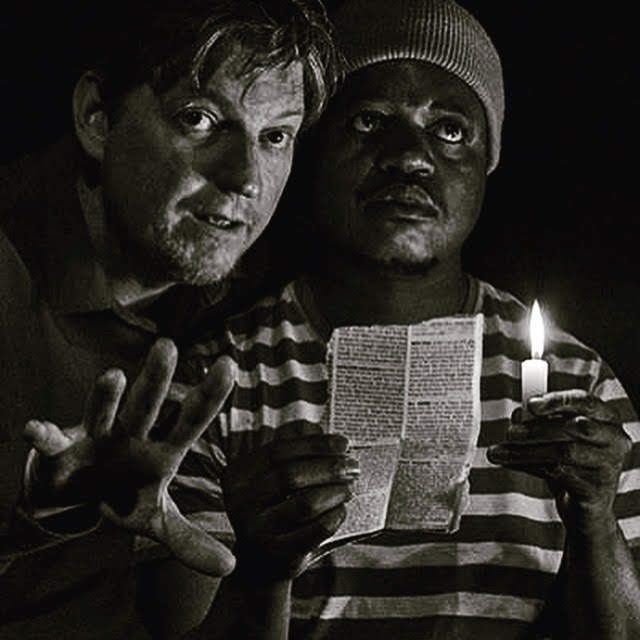 Black-and-white close-up of Clayton Pasi Gunguwo holding a candle and a newspaper clipping beside his scene partner