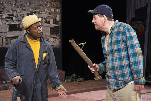 Clayton Pasi Gunguwo in coveralls and a yellow hat, facing a scene partner holding a pipe, in front of a township backdrop